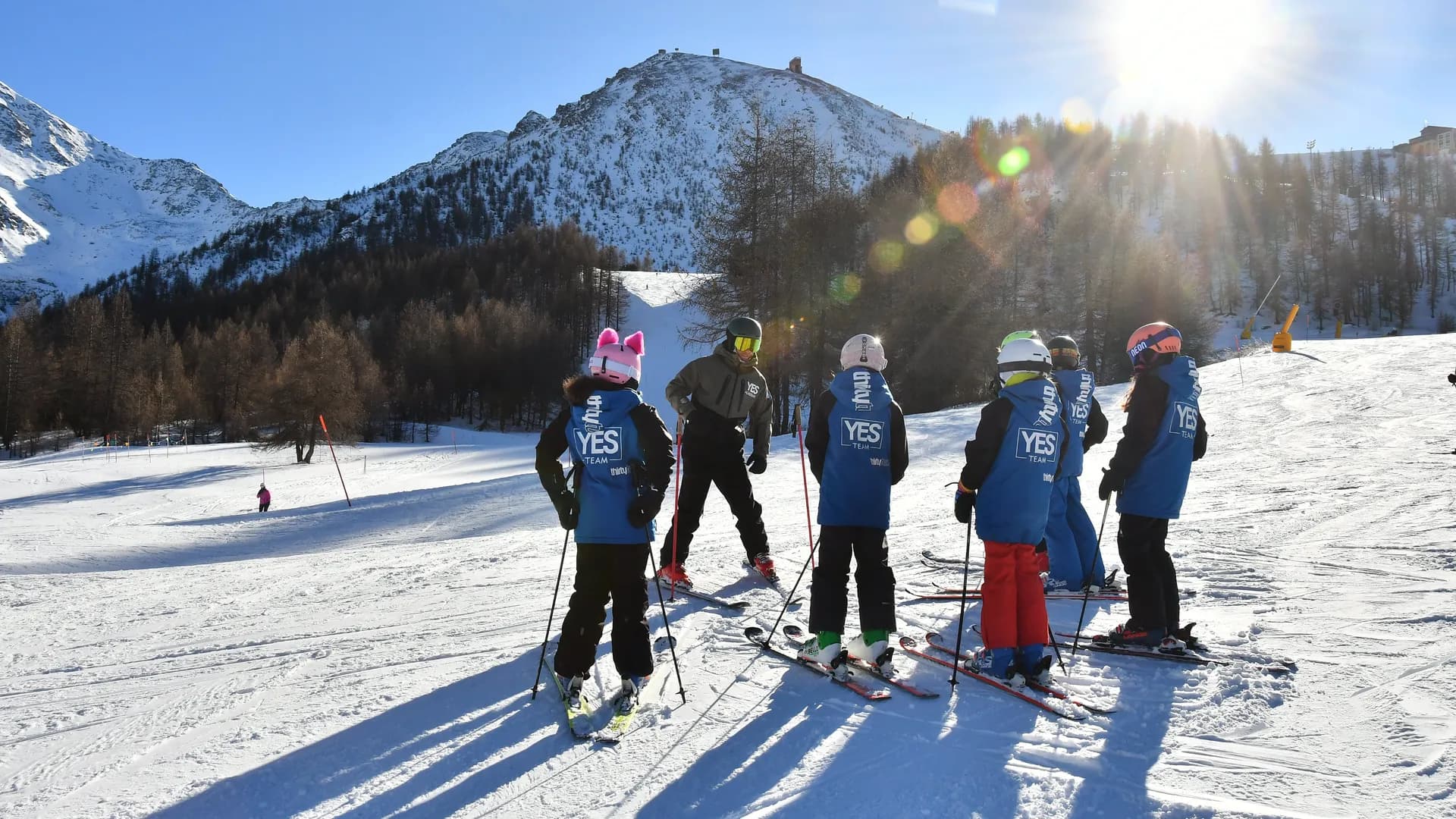 Il maestro spiega una curva al proprio gruppo di ragazzi di YES Team, in una giornata soleggiata e con le montagne che fanno da sfondo