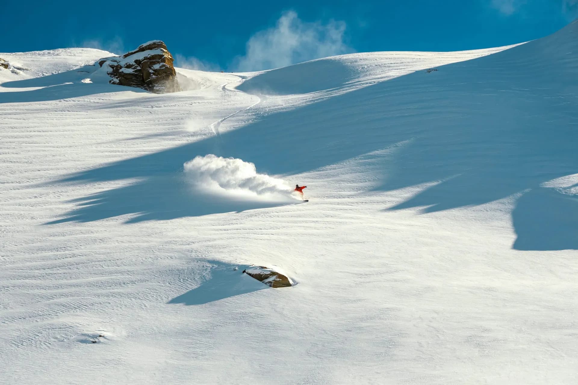 Rider in a red jacket riding fresh powder down a wide off-piste slope, throwing up a cloud of snow