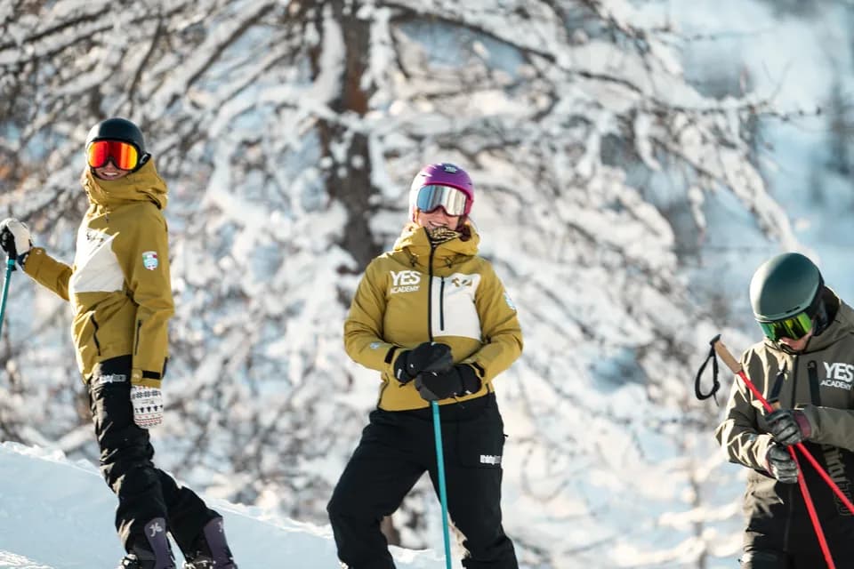 Trois moniteurs en veste YES Academy souriant sur la piste devant un arbre chargé de neige