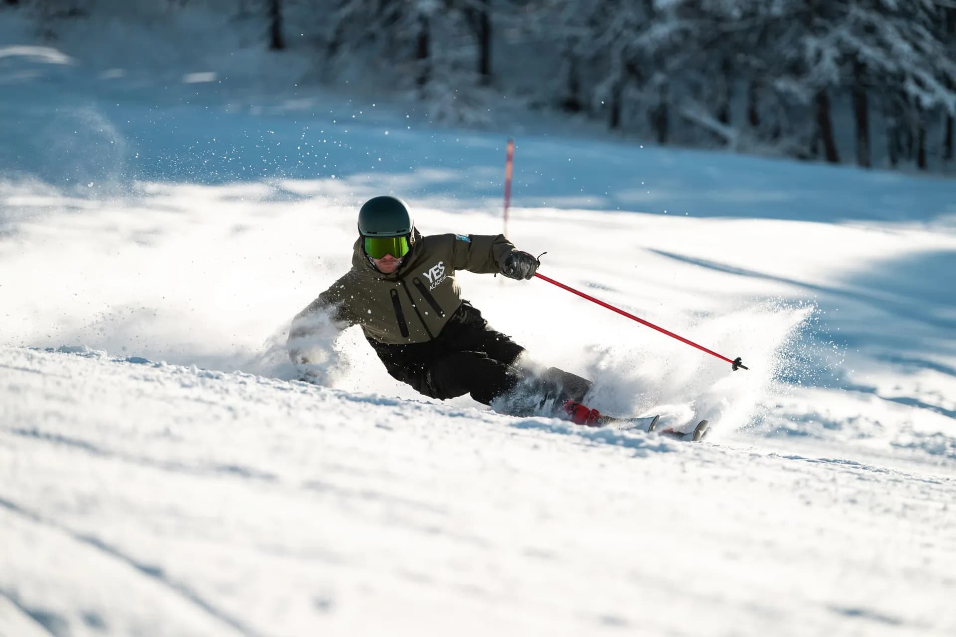 Skieur en veste YES Academy dans un virage serré, bâtons rouges et gerbe de neige, forêt enneigée au fond