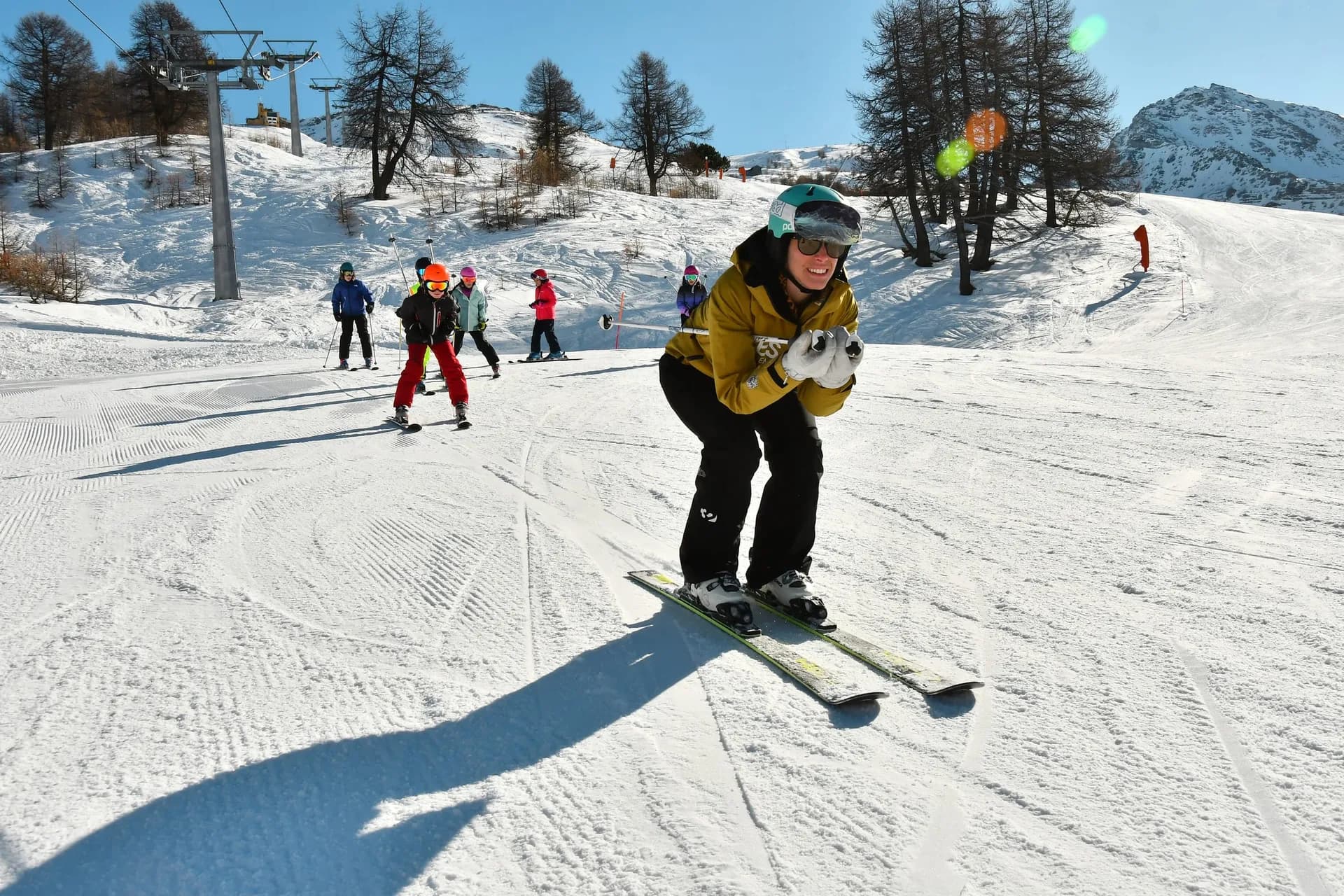 Ski instructor in a tuck leading a line of children down the piste, with a chairlift and larches behind