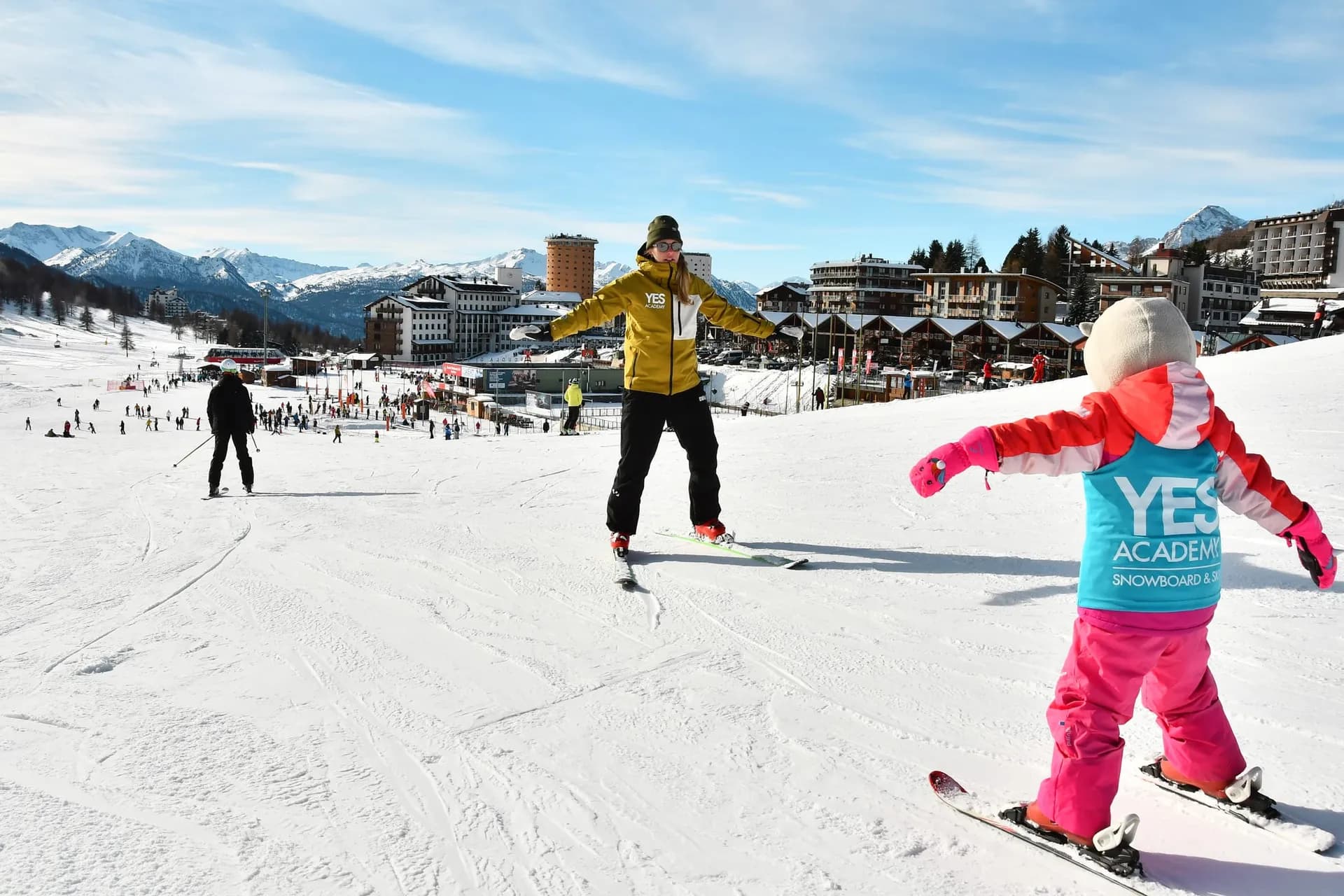Ski instructor with open arms guiding a small child on skis in a YES Academy bib, with Sestriere behind