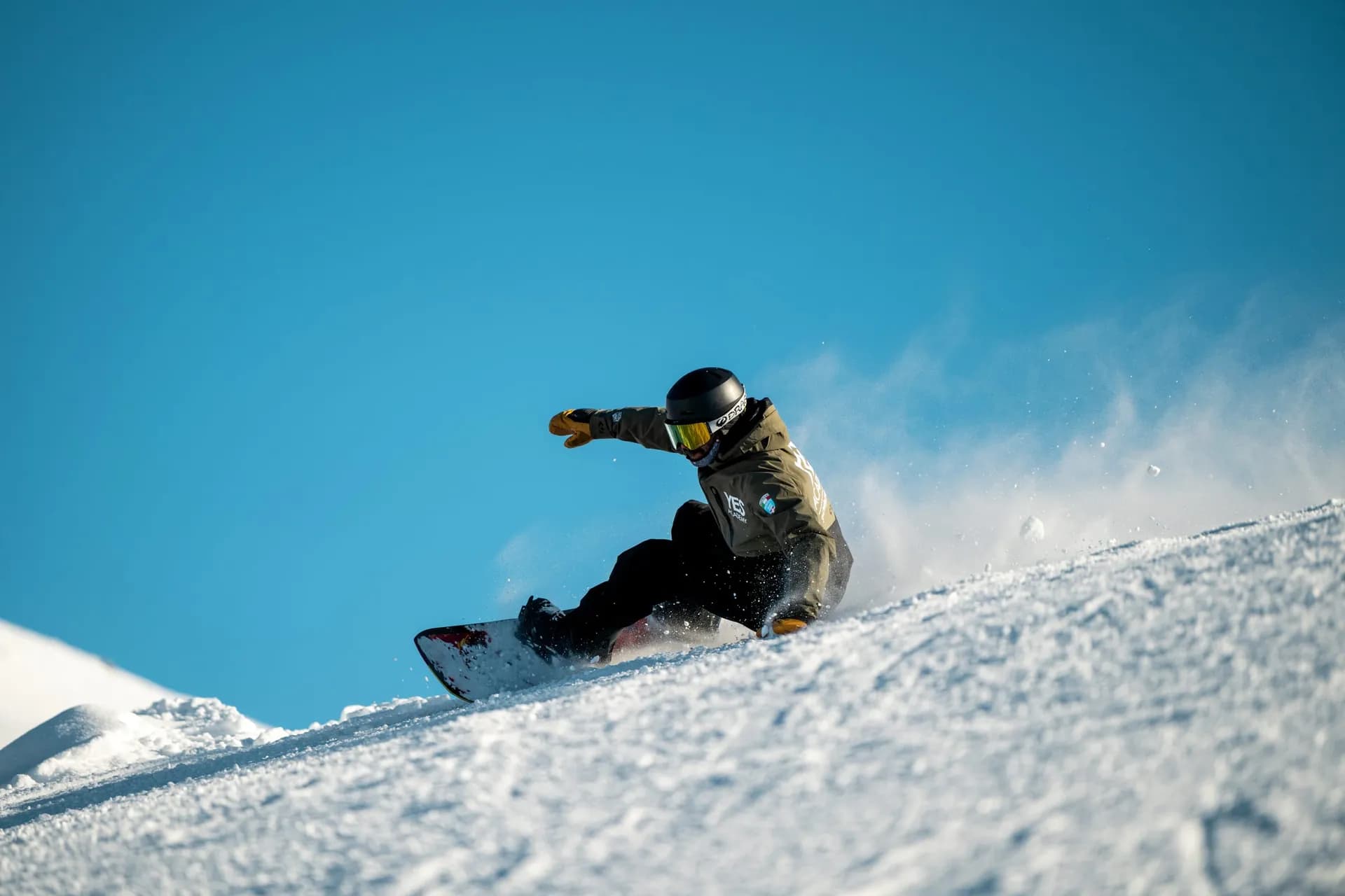 Snowboardeur en veste YES Academy en plein virage sur la piste, bras tendu, gerbe de neige sur fond de ciel bleu