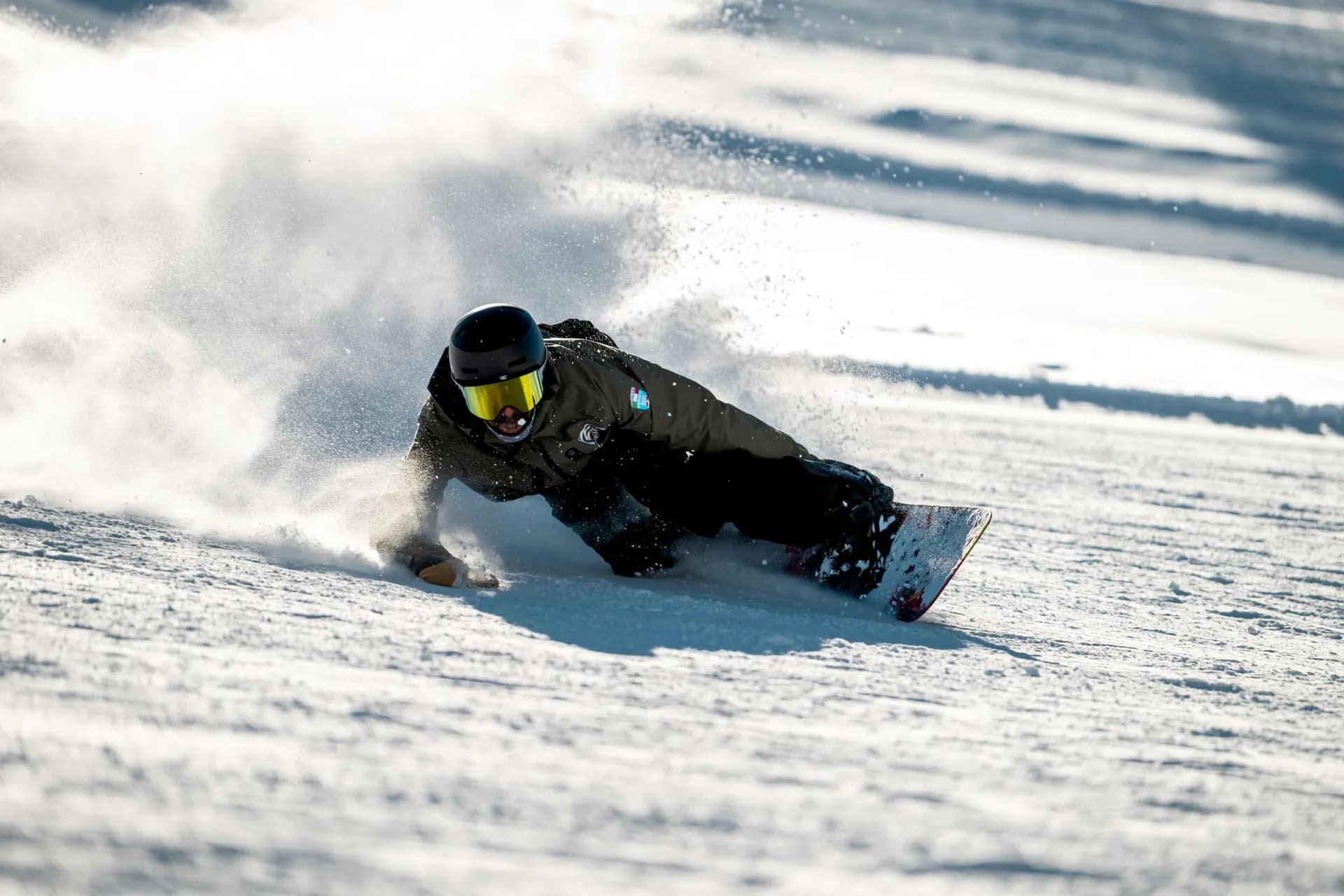 Snowboardeur YES Academy très incliné dans un virage, la main frôle la neige damée et soulève un nuage de neige