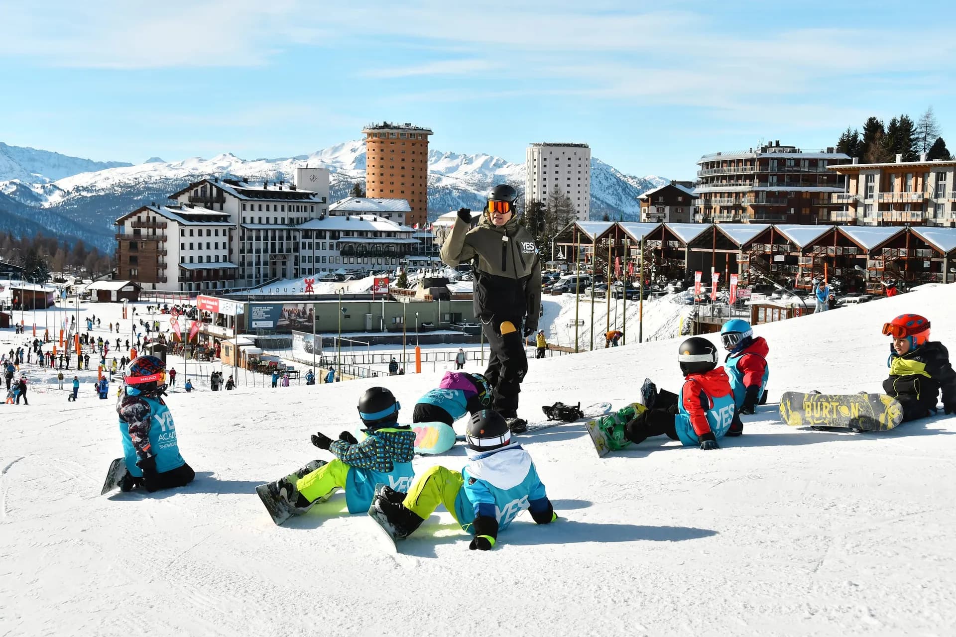 Snowboard instructor talking to seven children sitting in the snow with their boards, with Sestriere and the Alps behind