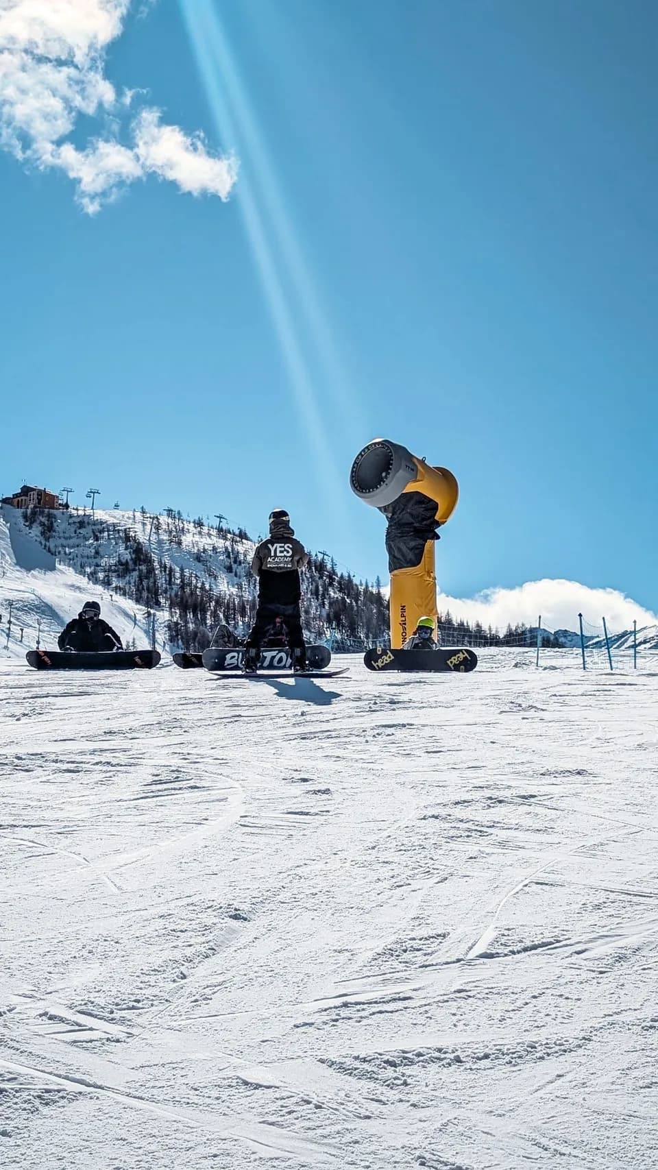 Moniteur de snowboard de dos face aux élèves assis avec leur planche, à côté d’un canon à neige jaune