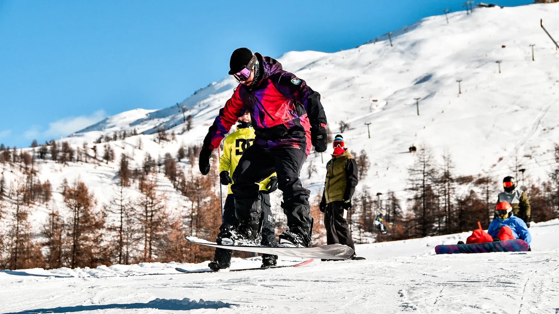 Snowboarder esegue un trick in pista in una giornata di sole, il resto del gruppo lo guarda. Sullo sfondo si vedono le montagne innevate.