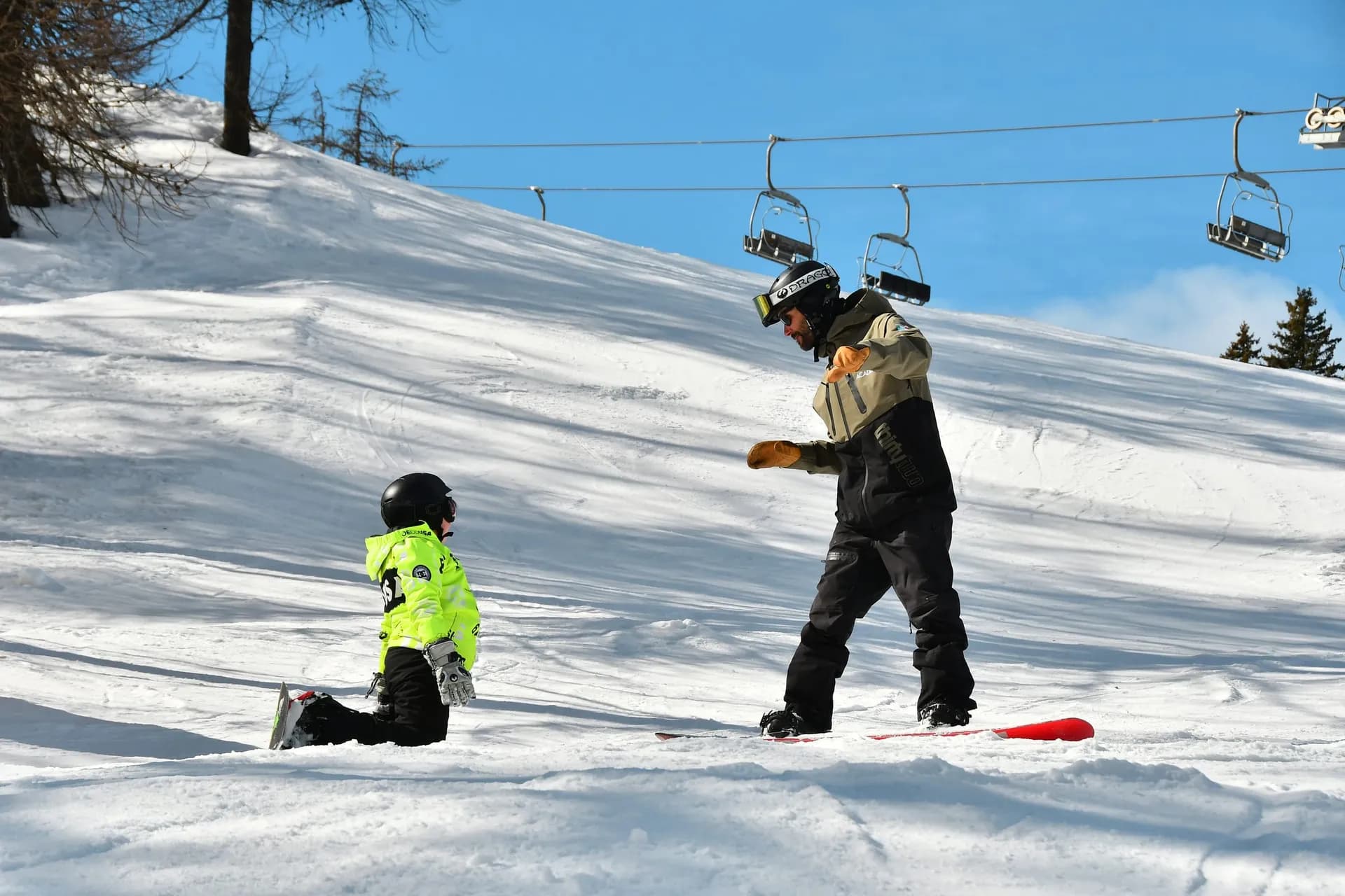 Snowboard instructor showing a move to a child sitting in the snow with a board, chairlift behind